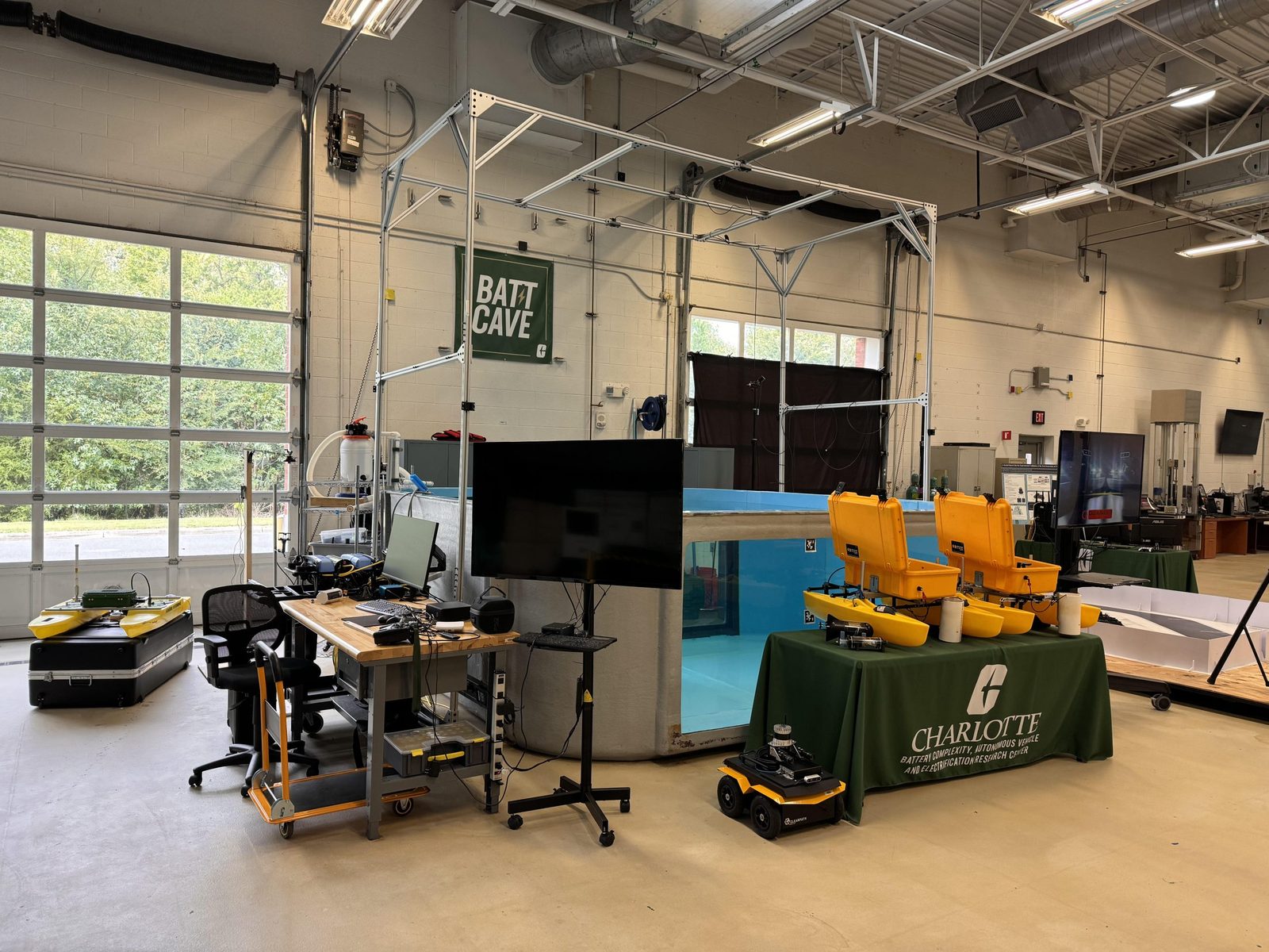 The BATT CAVE high bay, with the lab's water tank at centre, a bank of yellow seats behind it, ground robots and instrumentation carts in the foreground, and a tall roll-up door along the far wall.