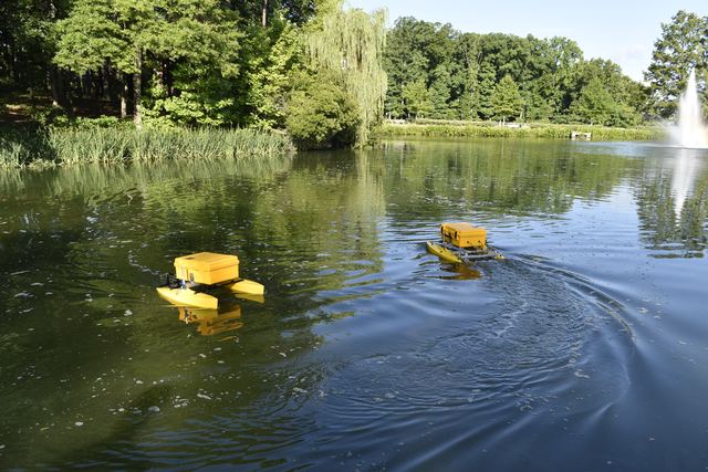 A yellow twin-hulled autonomous surface vessel underway on a pond, with fountains and a wooded bank behind it.