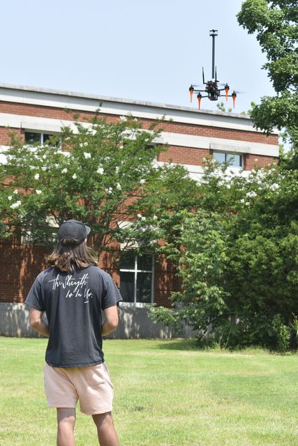 A person standing on a lawn beside a brick building, flying a quadrotor overhead.