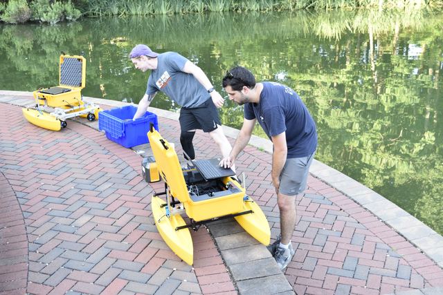 Three students on a brick walkway beside a pond, preparing a yellow twin-hulled autonomous surface vessel for launch.