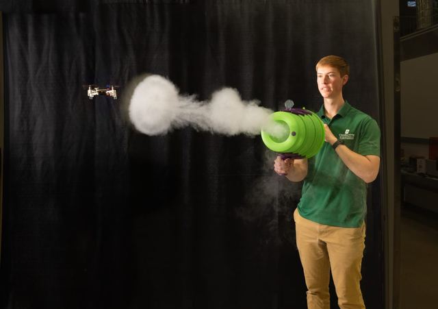 A student fires a vortex air cannon in a darkened lab, sending a visible smoke ring toward a small quadrotor.