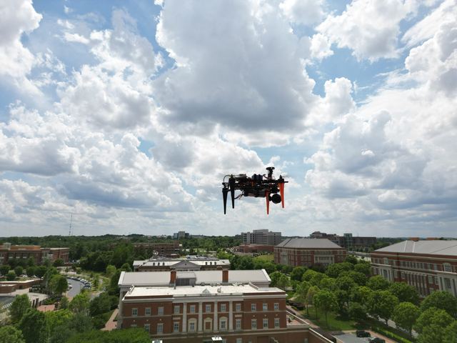 A quadrotor hovering high above the UNC Charlotte campus under cumulus clouds.