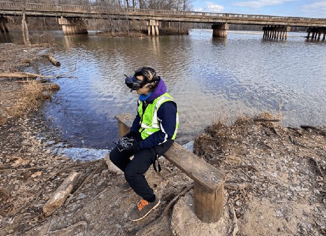 A researcher in a life vest sits on a log at a river bank operating a vehicle controller, with a railway trestle in the distance.