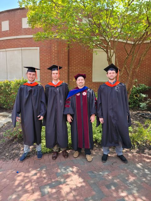 Graduates in academic regalia posed together outside a brick building.