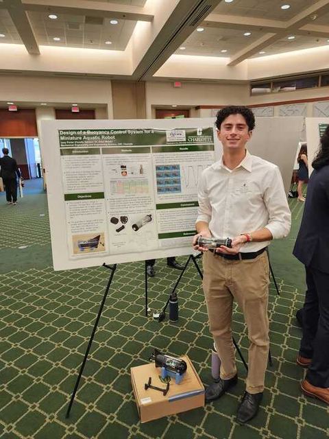 A student presenting a research poster at a conference while holding a small robot.