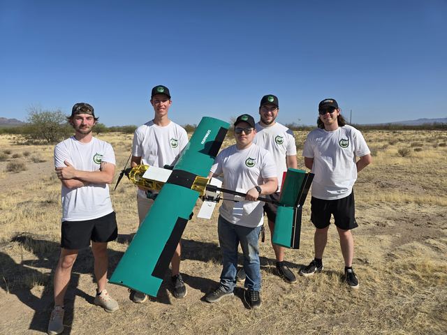 Five team members standing in an open desert test field, holding a large green and white fixed-wing aircraft.