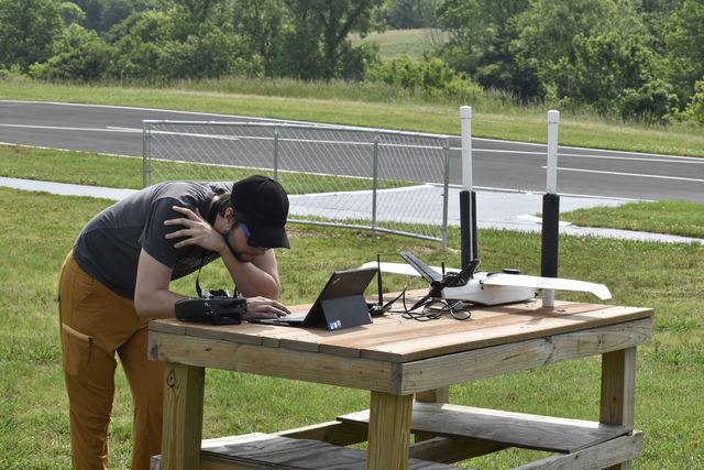 A researcher working at a laptop on an outdoor test table with antenna masts, beside a runway.