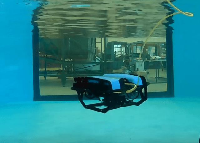 A small remotely operated underwater vehicle manoeuvring inside the lab water tank, seen through the acrylic viewport.