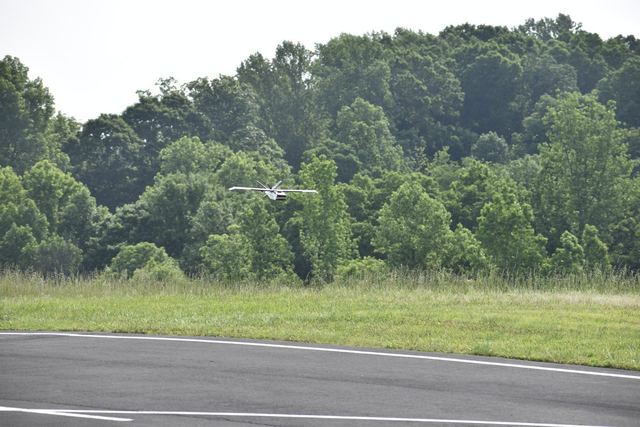 A fixed-wing UAV in flight low over a runway, with a tree line behind.