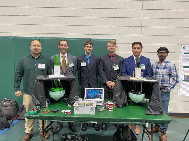 Six people standing behind a table displaying wave energy converter hardware at a demonstration event.