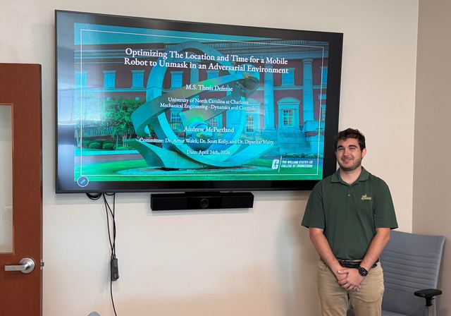 A student standing beside a wall-mounted display showing a presentation on optimising a mobile robot’s unmasking in an adversarial environment.
