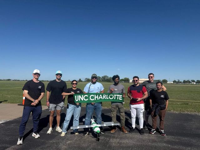 A team posed on a runway behind a UNC Charlotte banner with their aircraft.
