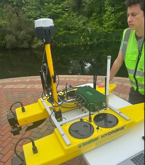 A yellow Seafloor Systems HYDRONE survey vessel on a brick plaza with its mast raised and a laptop set up beside it.