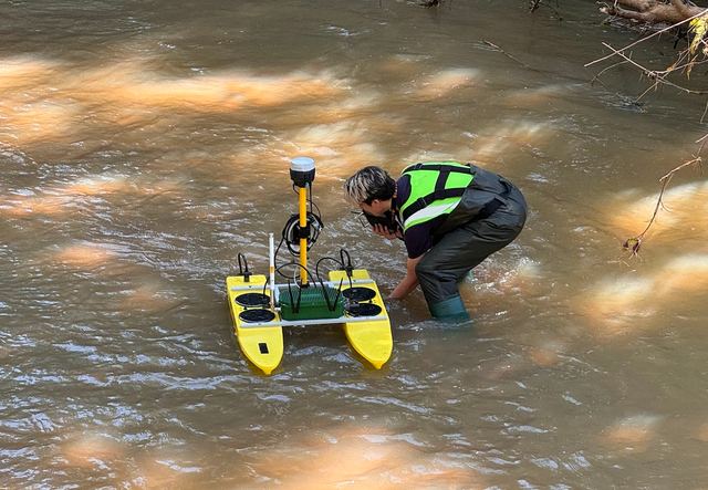 Two researchers in life vests crouch in shallow river water alongside a yellow autonomous surface vessel.