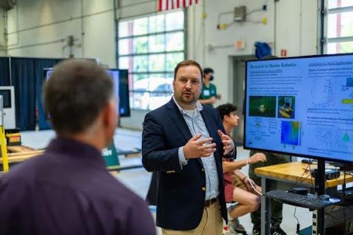 A researcher presenting to visitors in the high bay in front of a monitor showing research results.