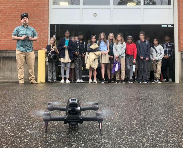 A researcher demonstrating a quadrotor to a group of schoolchildren outside a brick building.